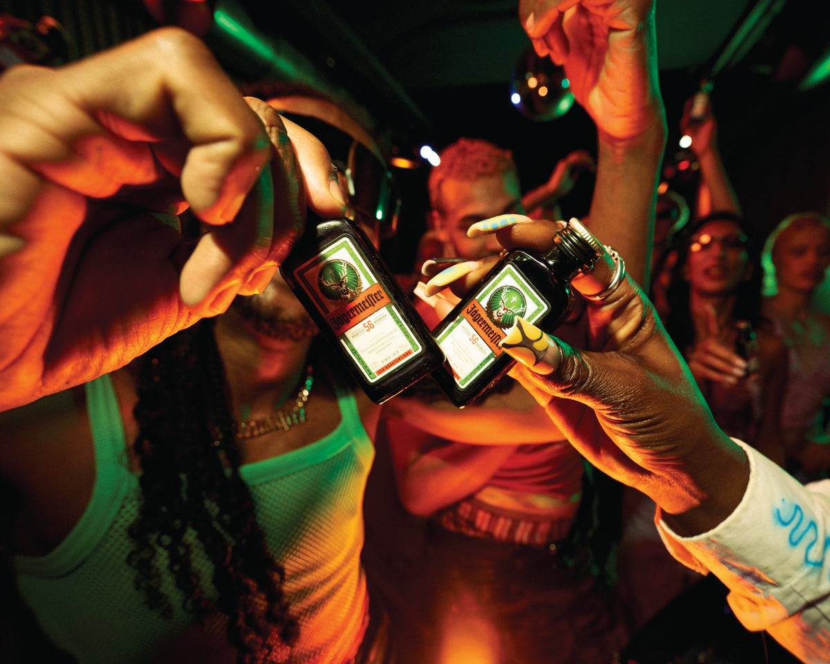 Two people holding up bottles of Jägermeister in a nightclub setting with green and orange lighting, with other people blurred in the background.