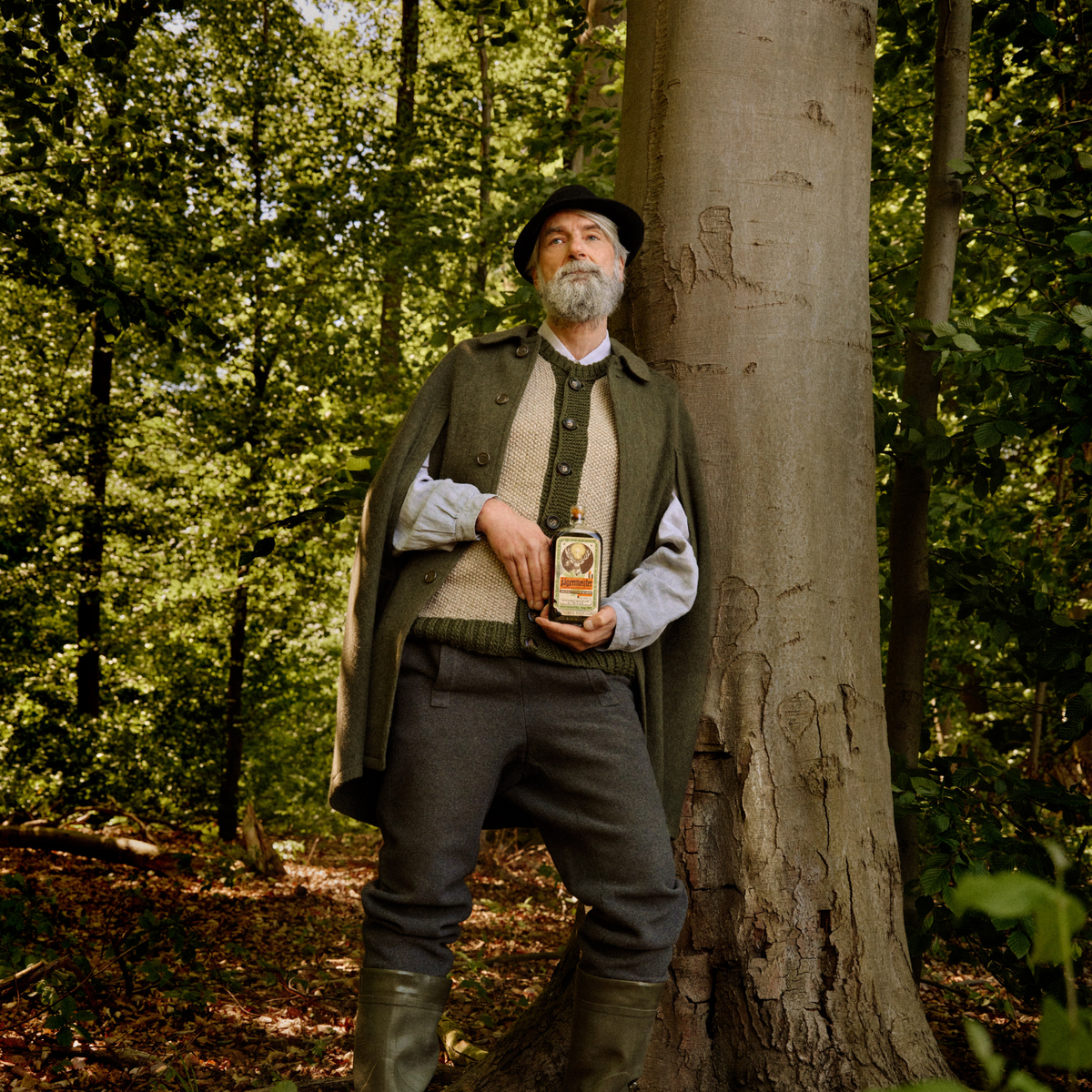 An older man with a gray beard stands in a forest next to a large tree trunk, wearing a dark green cape, beige sweater vest, light blue shirt, gray trousers, and green rubber boots while holding a retro-bottle of Jägermeister.