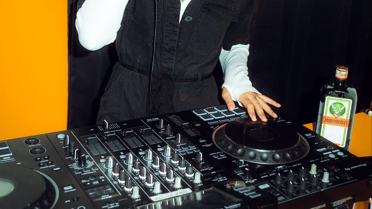 A DJ in a black vest and white shirt operating a black mixing console with turntables, with a bottle of Jägermeister on a table against an orange and black background.