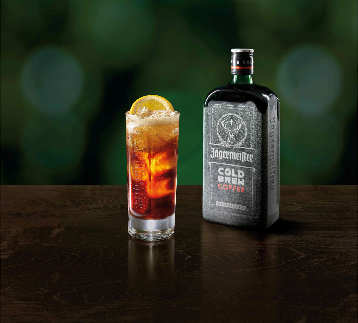 A bottle of Jagermeister Cold Brew Coffee stands next to a tall glass filled with icocktail drink and a lemon wheel garnish, set on a dark wooden surface against a blurred green background.