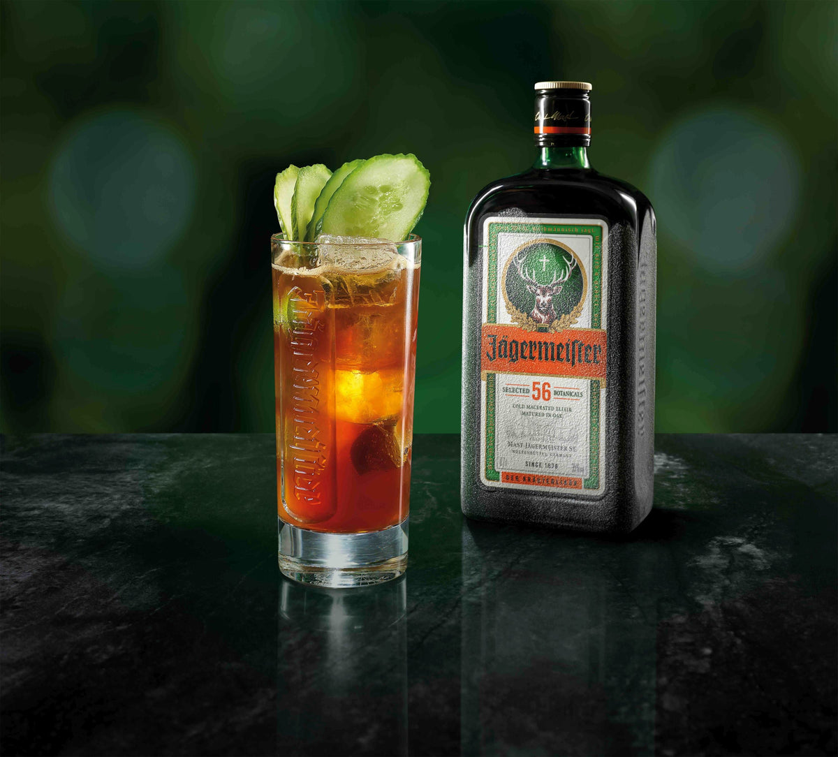 A Jägermeister bottle stands next to a tall glass cocktail filled with amber liquid and garnished with cucumber slices, on a dark reflective surface against a blurred green background.