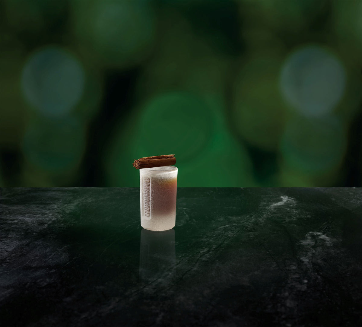 A small frosted shot glass with Jägermeisger and a cinnamon stick resting on top, set on a dark reflective surface against a blurred green bokeh background.