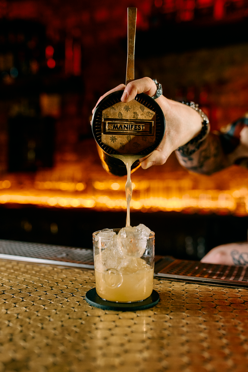 A hand pours a cocktail liquid from a metal strainer into a clear rocks glass filled with ice on a bar counter, with a warm, blurred bar background.