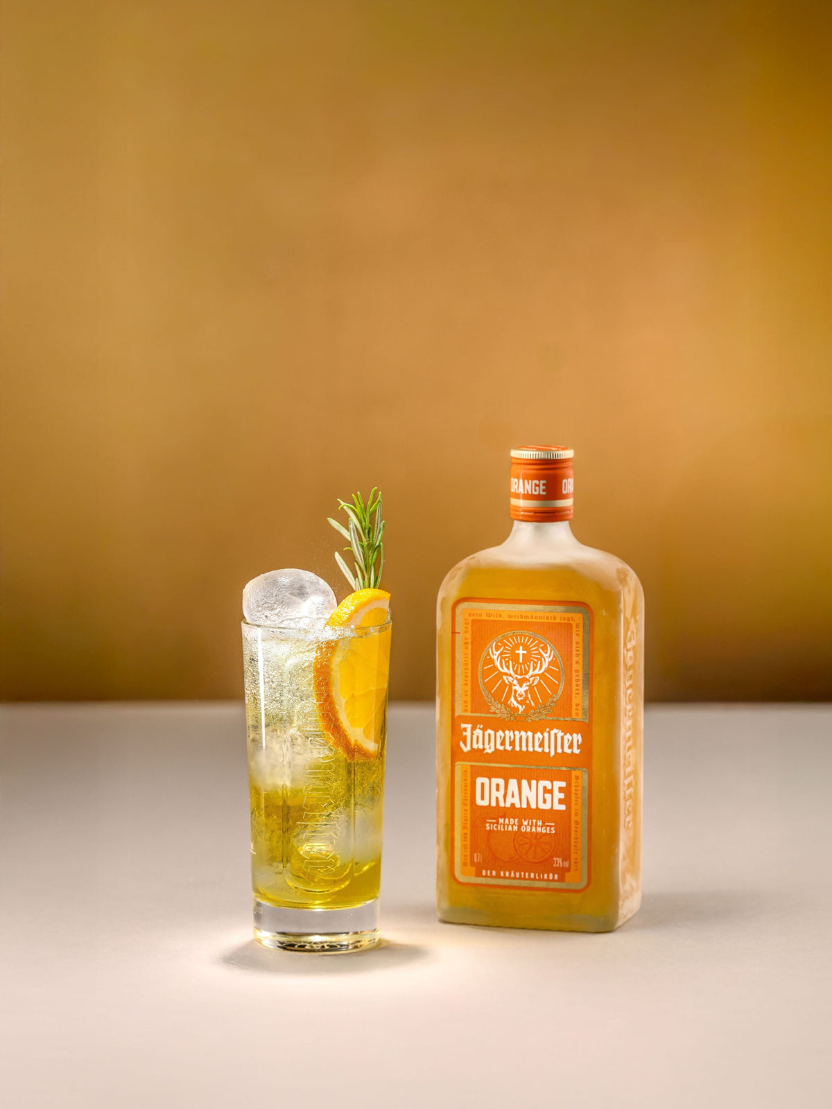 A bottle of Jägermeister Orange liqueur stands beside a highball glass filled with a golden drink, ice, an orange slice, and a sprig of rosemary against a warm gradient background.