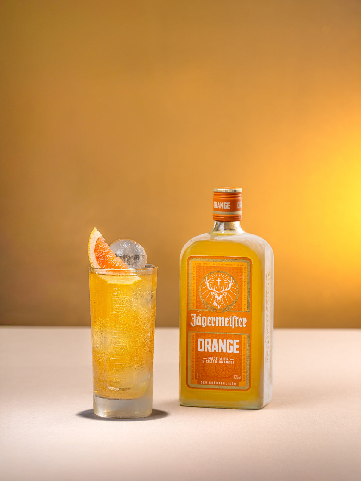 A bottle of Jögermeister Orange liqueur stands beside a tall glass filled with the same orange liquid, garnished with an orange slice and a round ice piece, against a warm golden studio background.