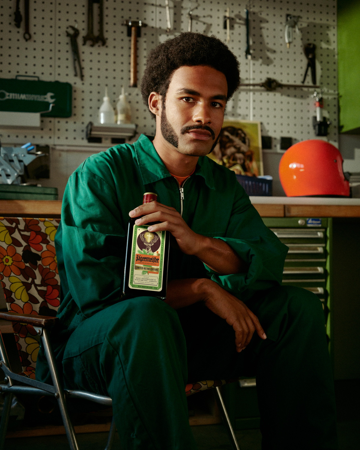 A man in a green mechanic jumpsuit sits in a garage holding a Jägermeister retro-bottle.