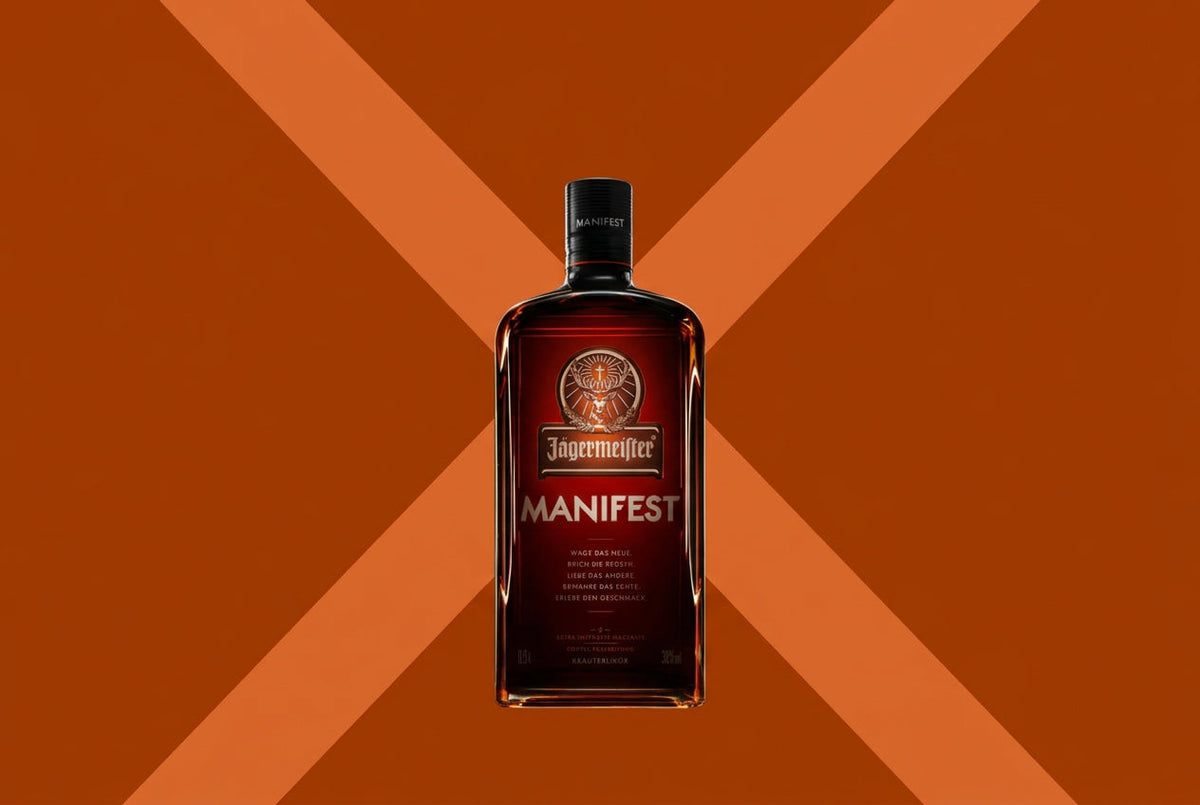 A bottle of Jagermeier Manifest liquor centered against a solid orange background with a large dark orange X behind it.
