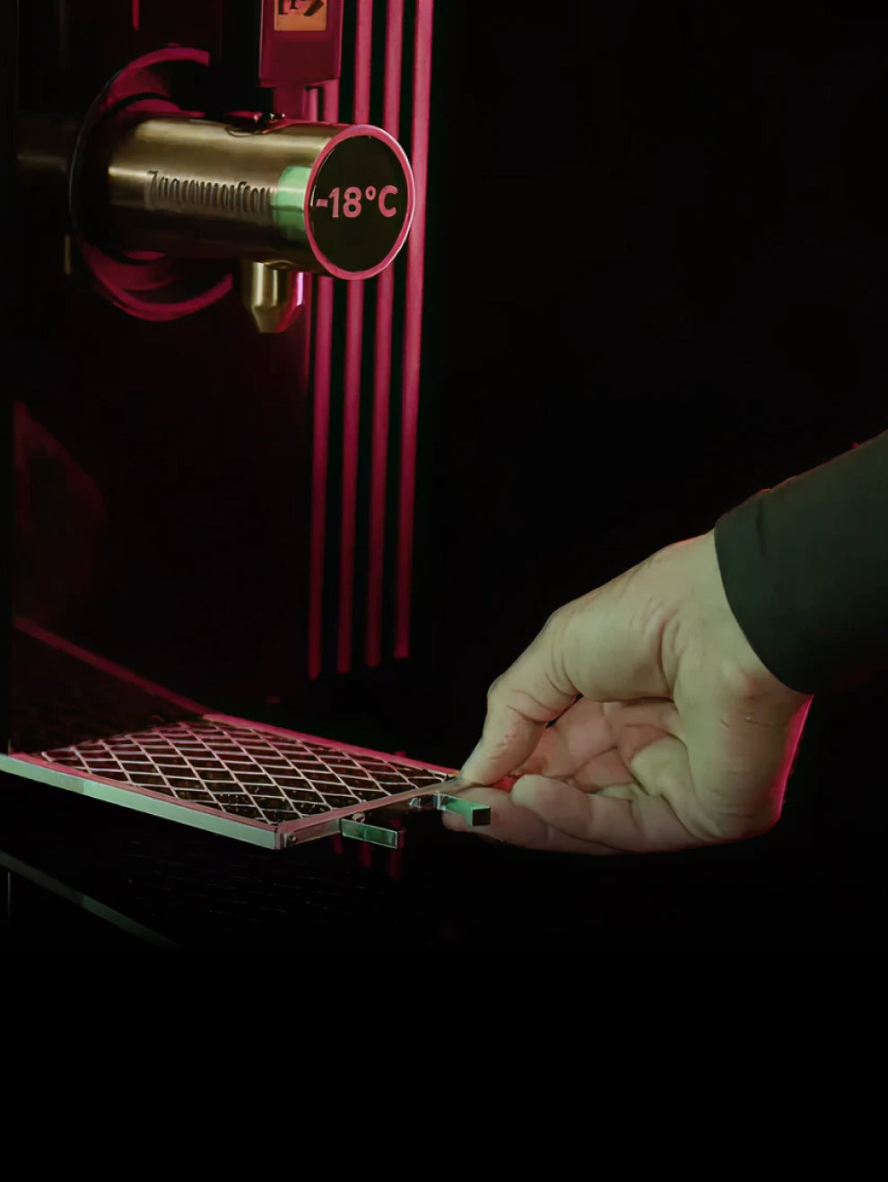 A hand pulls out a small metal tray from a dark green Jägermeister dispenser, which has a thermometer showing -18°C.